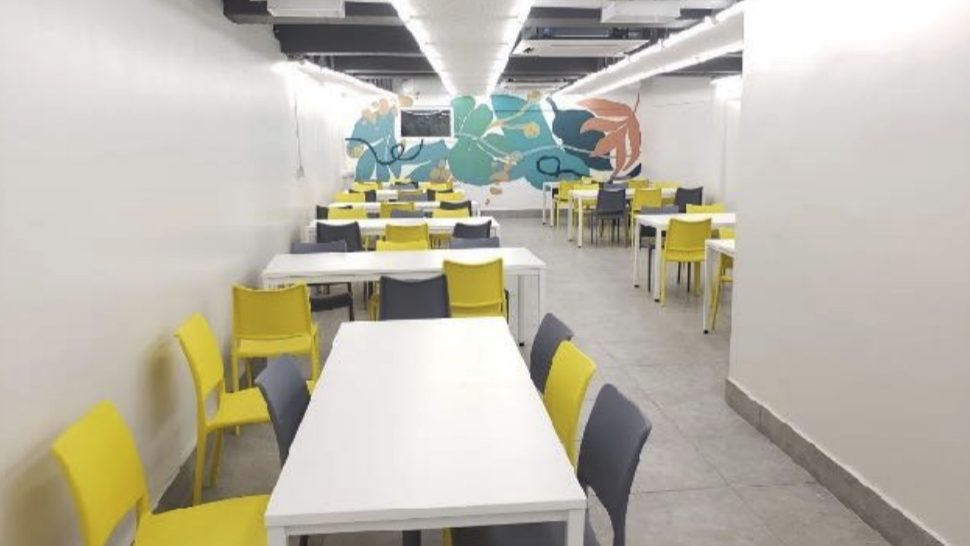Comedor para colaboradores con mesas blancas, sillas amarillas y grises, y un mural de hojas al fondo