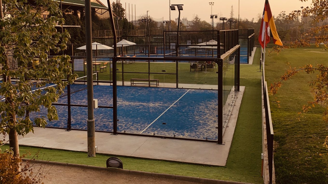 Cancha de pádel de piso azul con estructura metálica y cristal, rodeada de pasto, árboles y quitasoles